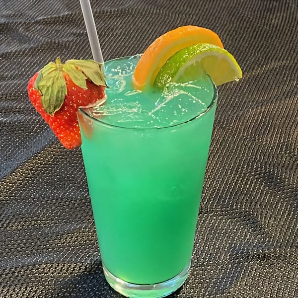 Bright green cocktail with a straw, garnished with a strawberry, orange slice, and lime.