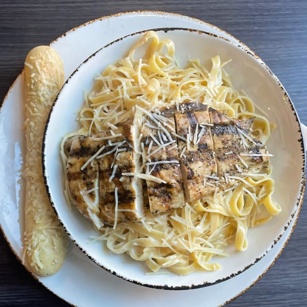 Grilled chicken Alfredo pasta with shredded cheese and a cheesy breadstick on a white plate.