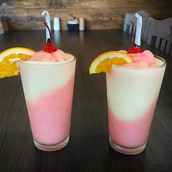 Two pink and white frozen drinks with orange slices and cherries on a dark wooden table.